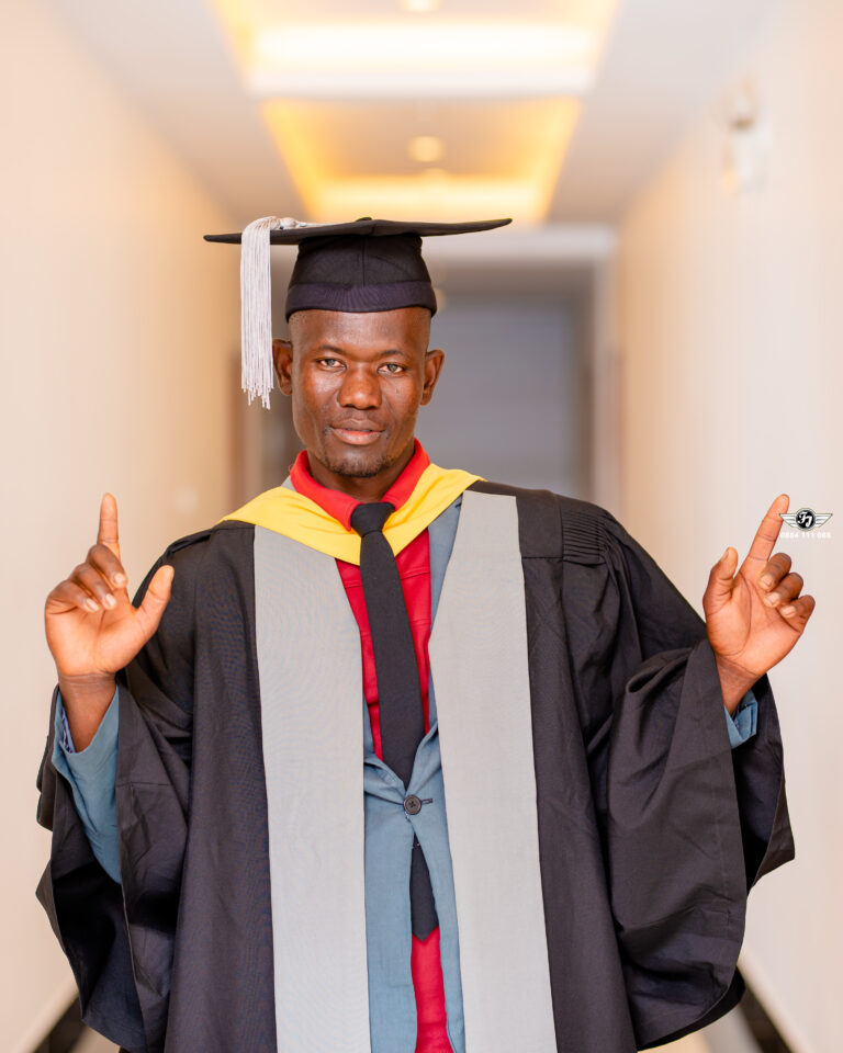 Sponsored student Alick Nathan poses in a white hallway. He is wearing a graduation robe and cap.