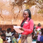 An African woman in a red jacket and yellow skirt stands in a crowd of women, reading to them from a paper.
