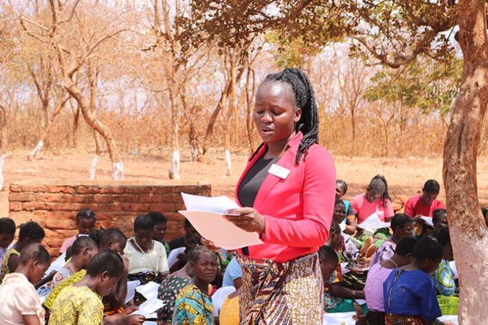 An African woman in a red jacket and yellow skirt stands in a crowd of women, reading to them from a paper.