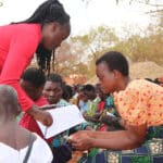 An African woman with braided hair and a red jacket bends over to help another African woman with short hair and an orange shirt fill out a piece of paper.