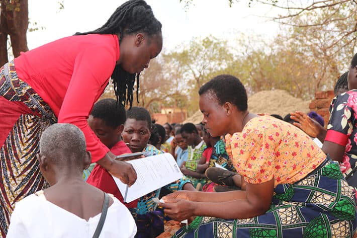 An African woman with braided hair and a red jacket bends over to help another African woman with short hair and an orange shirt fill out a piece of paper.