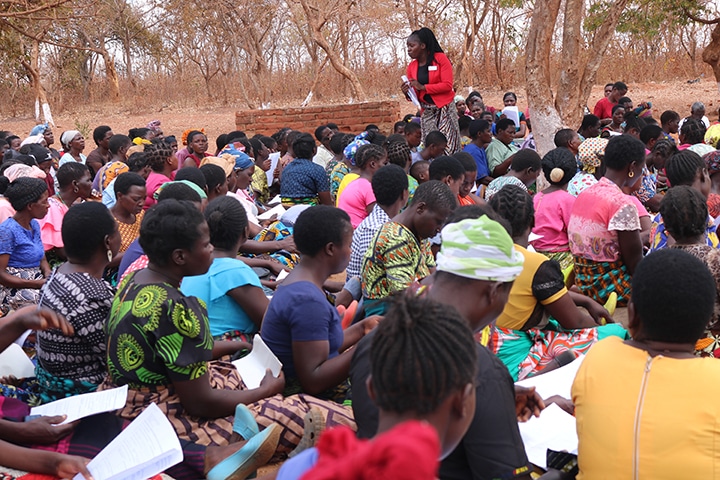 A woman in a red jacket stands up, talking to a crowd of women sitting down outdoors.