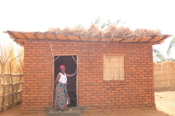 An African Woman in a white shirt and colorful skirt stands in the doorway of a small red brick building with a thatched grass roof.