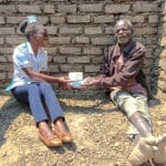A nurse and a patient are sitting on the ground in front of a brick wall. The nurse is handing the male patient a box of medical supplies.