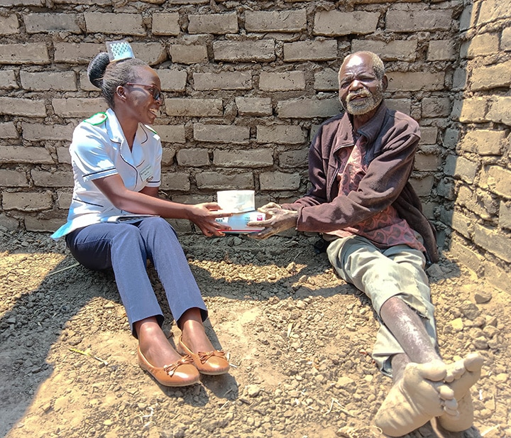 A nurse and a patient are sitting on the ground in front of a brick wall. The nurse is handing the male patient a box of medical supplies.