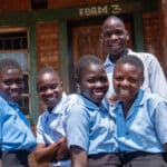 A group of five African students smiles together, wearing light blue shirts and dark blue pants and skirts. There are three girls and two boys.
