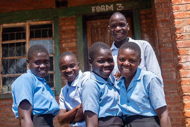 A group of five African students smiles together, wearing light blue shirts and dark blue pants and skirts. There are three girls and two boys.