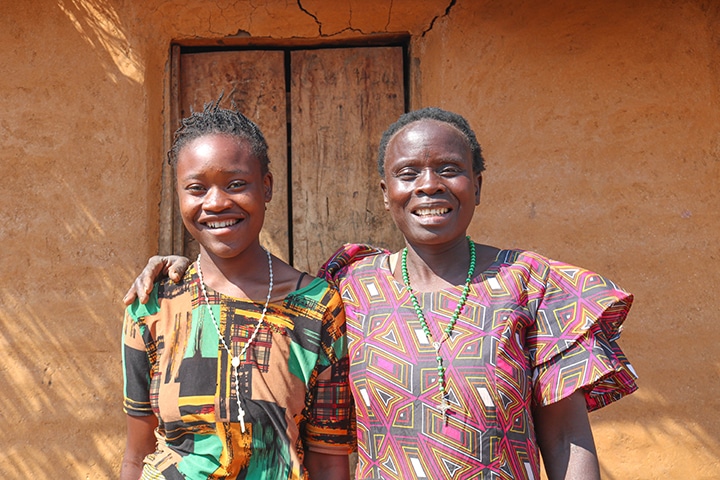 An African teenage girl and her mother stand together in front of their home, smiling.