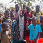 African children of various ages stand around a water well. They are smiling and giving Thumbs Up signs.