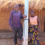 An African man and his African wife stand in front of a mud brick hut with a roll of metal sheeting for their roof.