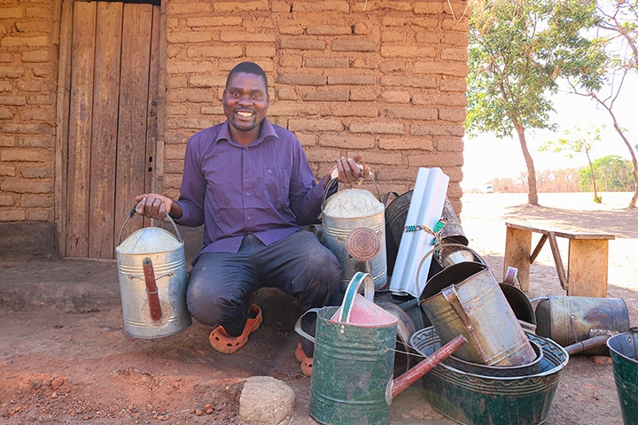 An African man squats in front of a mud brick hut with a pile of tin watering cans.