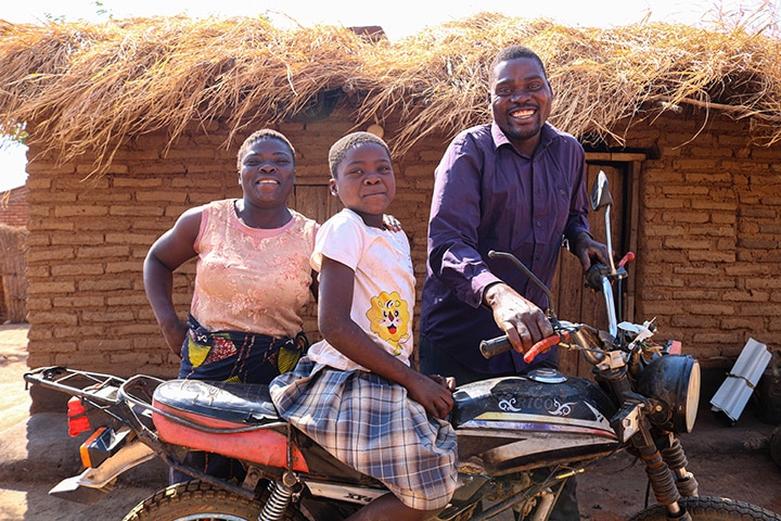 A woman, child and man stand with a parked motorbike in front of a mud brick hut in Africa.