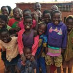 A small crowd of African children smile together in a group. They are wearing colorful, tattered clothing and standing in a dirt yard in front of a mud brick building.