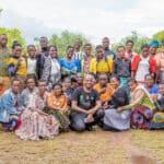 A group of African men and women stand together outdoors in front of trees, smiling and throwing up peace signs. They are dressed in brightly colored clothing, and some are wearing colorful headdresses. The ground is wet from the recent rains.