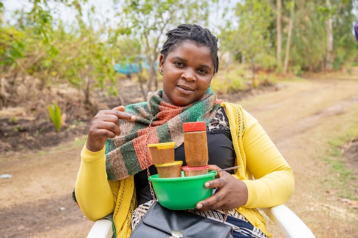 An African woman smiles as she holds a jar of tomato sauce that she has made.