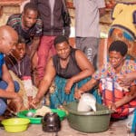 Several African women sit together mixing ingredients in colorful bowls.
