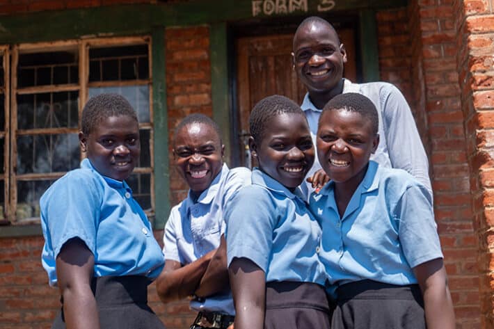 Five students stand together smiling and laughing. They are high school age, African, with dark skin and short cropped hair. They are wearing light blue shirts and dark blue or black pants or skirts. There are three girls and two boys.