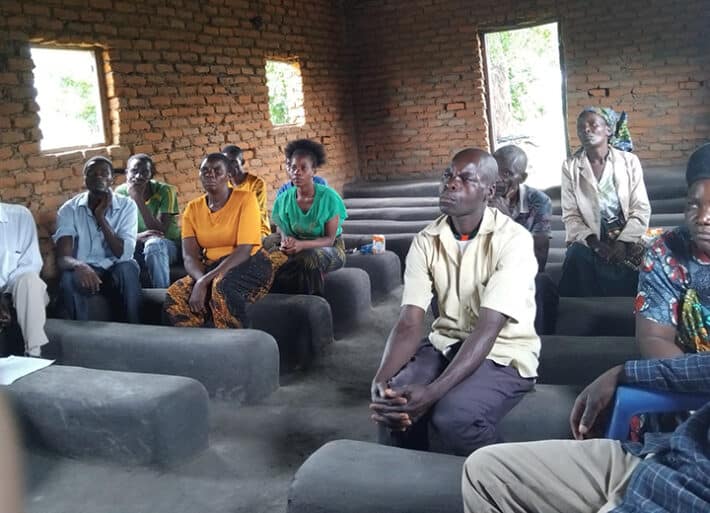 Several people sit on concrete benches in a brick building. They are Africa, wearing old clothes and have serious looks on their faces. They are attentively listening to the speaker.