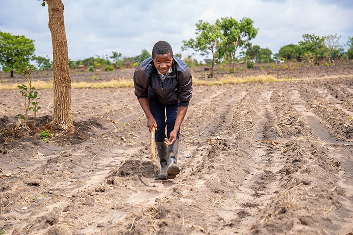 A young African person stands in a muddy field during planting time. They are leaning over, in the midst of planting seeds in the wet dirt.