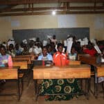 A classroom full of students smile and raise their papers in the air under the glow of solar powered lighting.