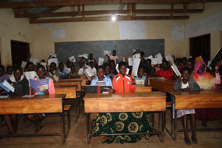 A classroom full of students smile and raise their papers in the air under the glow of solar powered lighting.