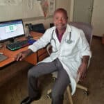 An African male clinician sits in a chair in front of a computer. He is wearing a white lab coat, grey slacks, a maroon shirt, and has a stethoscope around his neck.