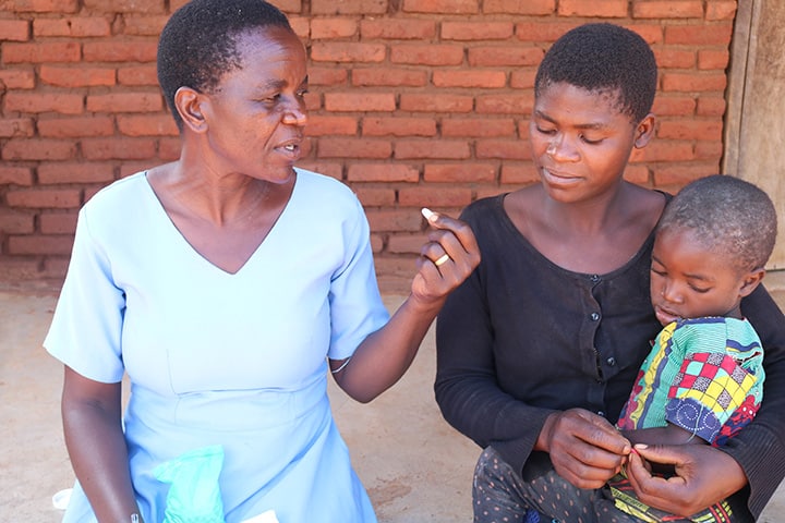 A nurse, wearing a blue dress, explains a medical supplement to a mother, who is holding her child in her arms.