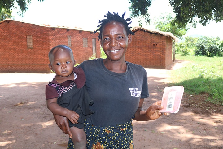 An African Mother holds her baby on her hip. She is smiling and holding a paper pamphlet. They are outdoors and the sun is shining.