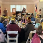 A crowd of people sit at various tables in a nicely appointed room in the sunlight. A man and woman are on stage, presenting a program.
