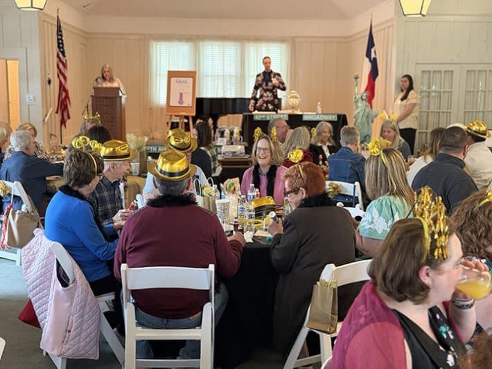 A crowd of people sit at various tables in a nicely appointed room in the sunlight. A man and woman are on stage, presenting a program.