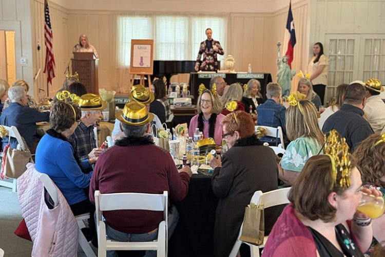 A crowd of people sit at various tables in a nicely appointed room in the sunlight. A man and woman are on stage, presenting a program.