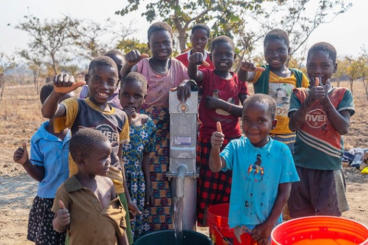 African children of various ages stand around a water well. They are smiling and giving Thumbs Up signs.