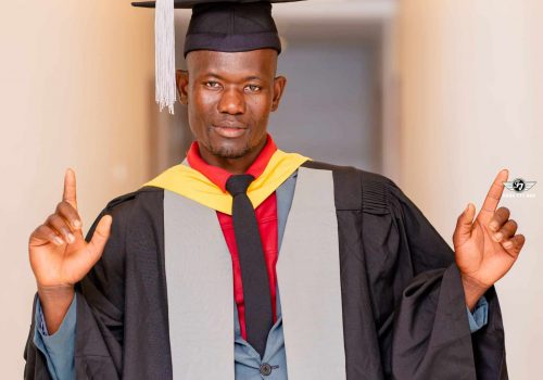 Sponsored student Alick Nathan poses in a white hallway. He is wearing a graduation robe and cap.
