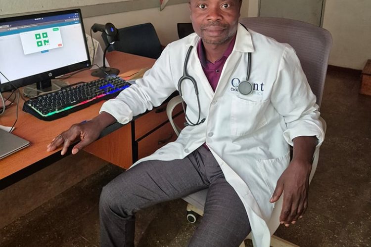 An African male clinician sits in a chair in front of a computer. He is wearing a white lab coat, grey slacks, a maroon shirt, and has a stethoscope around his neck.