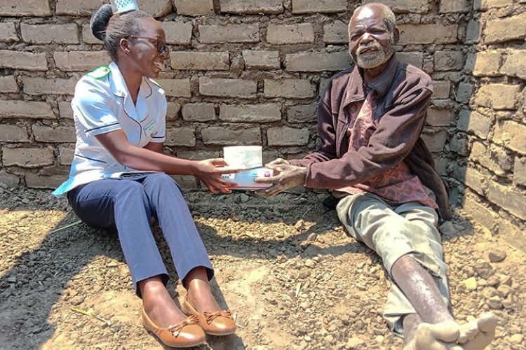 A nurse and a patient are sitting on the ground in front of a brick wall. The nurse is handing the male patient a box of medical supplies.