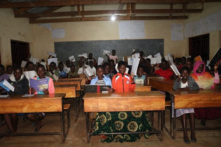 A classroom full of students smile and raise their papers in the air under the glow of solar powered lighting.