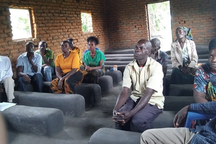 Several people sit on concrete benches in a brick building. They are Africa, wearing old clothes and have serious looks on their faces. They are attentively listening to the speaker.